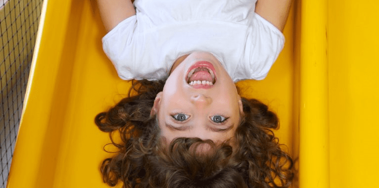 8 year old girl upside down on yellow slide with mouth open and blue eyes